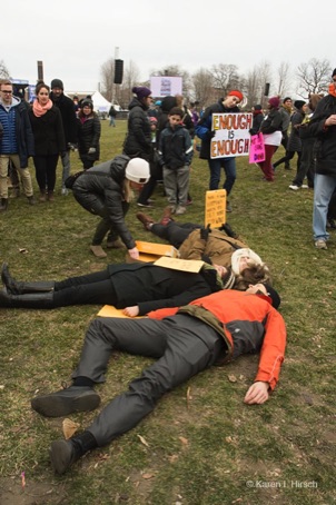 3 students play dead. In background, woman holds sign Enough is Enough